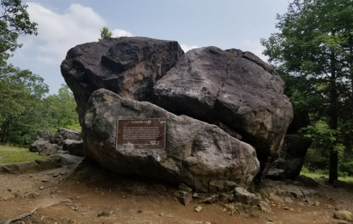 A large boulder with a plaque stands in a grassy area, surrounded by trees under a partly cloudy sky.
