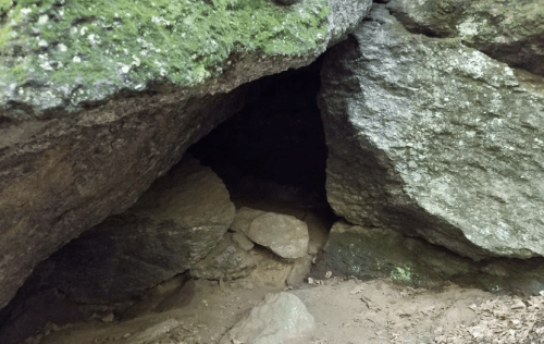 A dark cave entrance surrounded by large, moss-covered rocks and scattered stones on the ground.