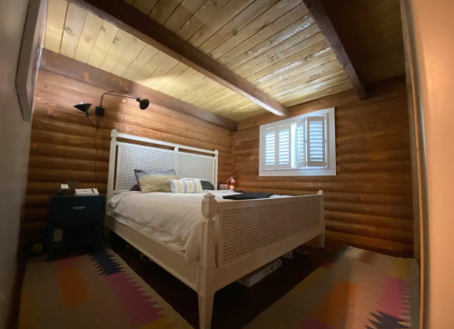 Cozy bedroom with a white bed, wooden walls, and a colorful rug, featuring a window with shutters and a bedside lamp.