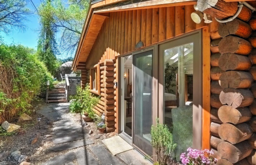A rustic log cabin with large glass doors, surrounded by greenery and flowers, under a clear blue sky.