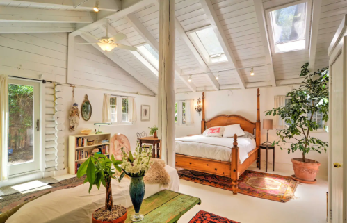 Bright, airy bedroom with wooden bed, large windows, plants, and cozy decor in a rustic setting.