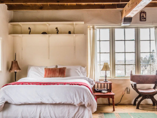 Cozy bedroom with a white bed, wooden shelves, and large windows, featuring warm lighting and rustic decor.