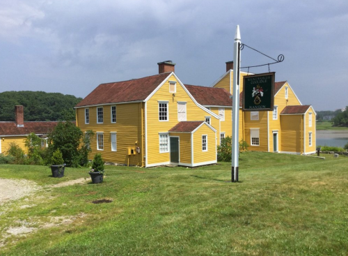 A yellow historic mansion with a sign, surrounded by green grass and trees under a cloudy sky.