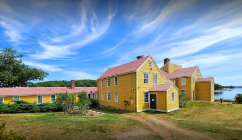 A bright yellow house with a red roof, surrounded by greenery and a view of the water under a blue sky.