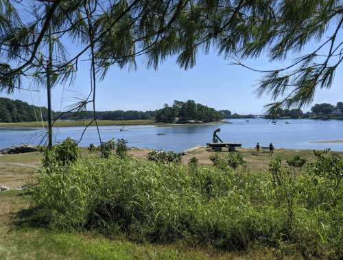 A serene waterfront scene with boats, a sculpture, and lush greenery under a clear blue sky.