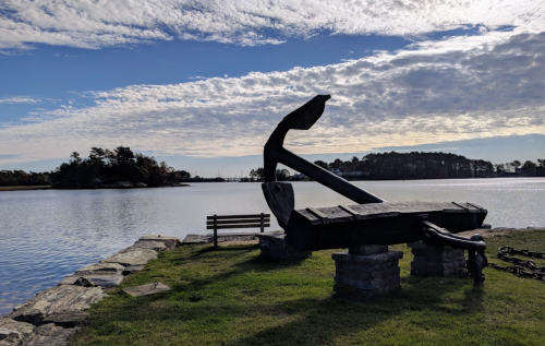 A large anchor sits on a stone pier by a calm waterway, with a bench and trees in the background under a cloudy sky.