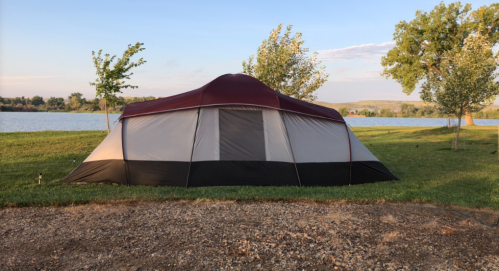 A large tent set up on grassy land near a calm lake, with trees in the background and a clear sky.