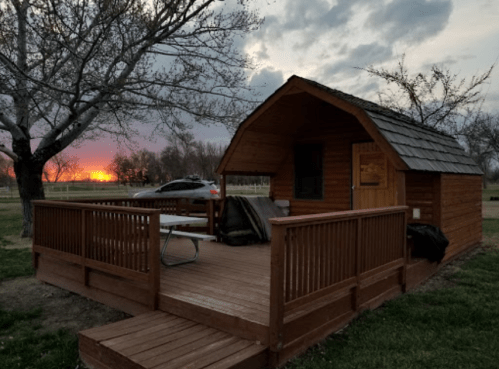 A cozy wooden cabin with a deck, surrounded by trees, against a sunset sky.