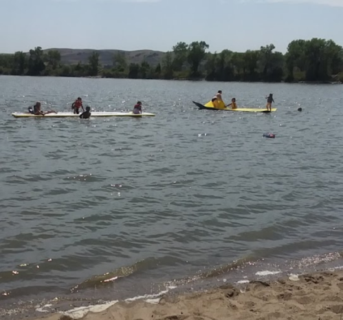 People paddleboarding and kayaking on a calm lake, with a sandy beach in the foreground and trees in the background.