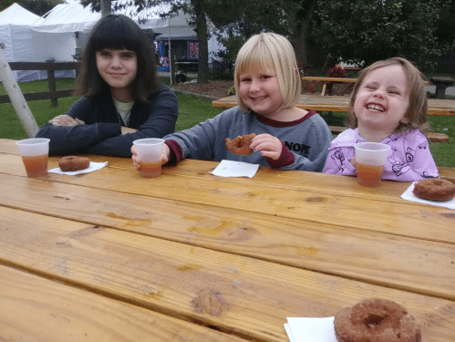 Three children sit at a wooden table, enjoying donuts and drinks, smiling in a park setting.