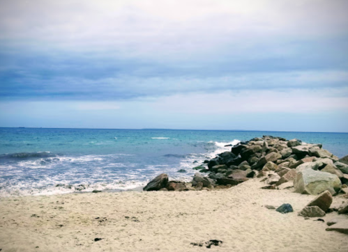 A rocky shoreline meets a calm sea under a cloudy sky, with gentle waves lapping at the sandy beach.