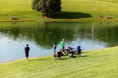 Two people fishing by a calm pond, with a picnic table and chairs nearby, surrounded by green grass and trees.