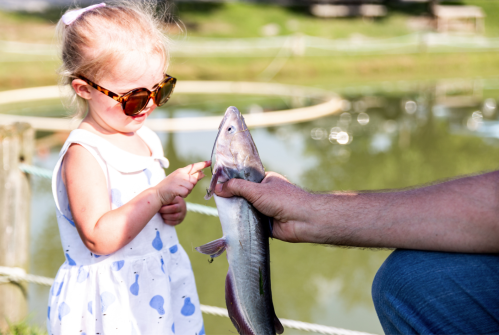 A young girl in sunglasses smiles as she points at a fish held by an adult near a pond.