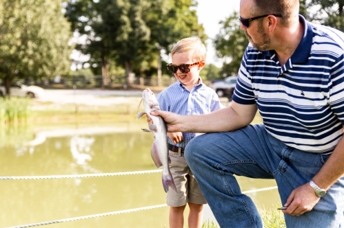 A man and a boy wearing sunglasses proudly hold a fish by a pond, with trees and a car in the background.