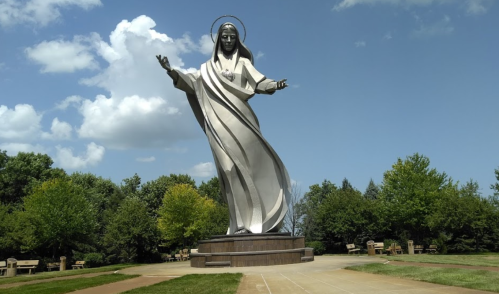 A large metallic statue of a figure with outstretched arms, surrounded by trees and a blue sky with clouds.