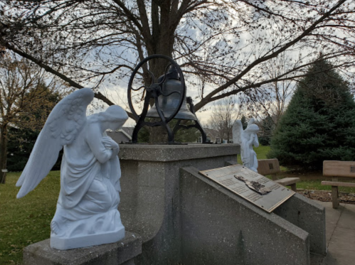 Two white angel statues flank a memorial with a bell and plaque, set under a tree in a peaceful park.