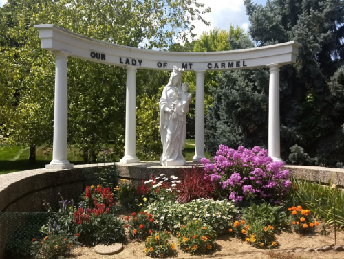 A statue of Our Lady of Mt. Carmel surrounded by colorful flowers and greenery under a blue sky.