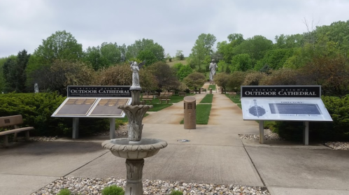 A serene outdoor cathedral with statues, benches, and informational signs amidst lush greenery and a cloudy sky.