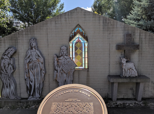 A stone wall features figures of saints and a stained glass window, with a cross and a lamb sculpture in front.