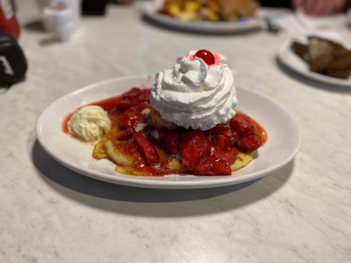 A plate of pancakes topped with strawberries, whipped cream, and a cherry, served with a scoop of ice cream.