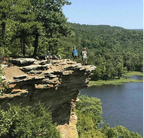 Three hikers stand on a rocky cliff overlooking a lush green landscape and a river below.