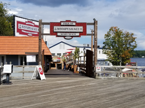 A wooden pier at Weirs Beach, Lake Winnipesaukee, with shops and a scenic view of the water and trees.