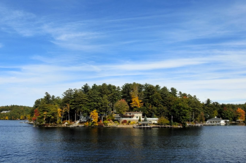 A serene island with a house surrounded by colorful autumn trees, set against a clear blue sky and calm water.