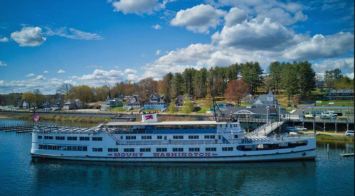 A large white steamboat named "Mount Washington" docked by a waterfront with trees and buildings in the background.