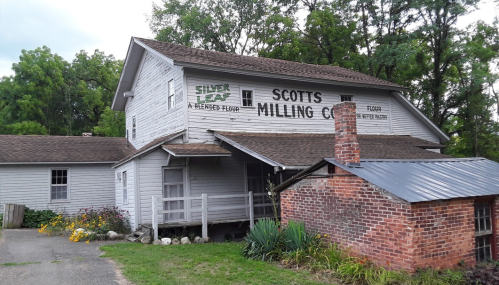 Historic building with "Scott's Milling Co." sign, featuring a brick chimney and surrounded by greenery.