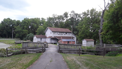 Historic building with a sign reading "Scott's Milling Co," surrounded by trees and a pathway leading to it.