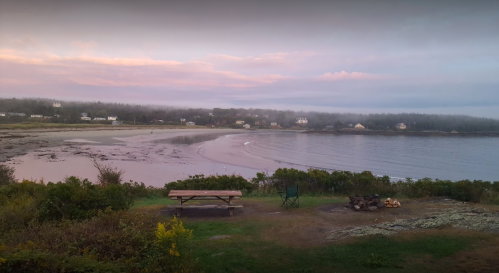 A serene beach at dawn, with gentle waves, misty hills, and a picnic area overlooking the water.