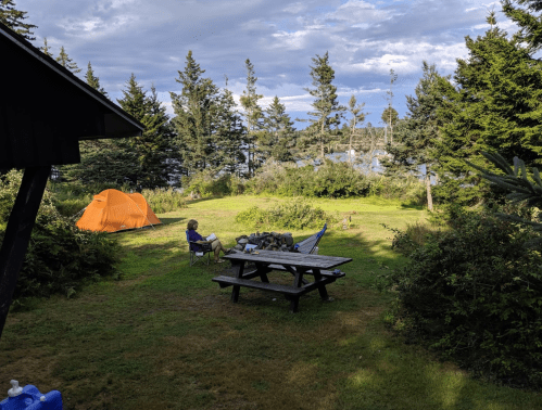 A campsite with a person sitting by a picnic table, an orange tent, and trees by a lake under a cloudy sky.