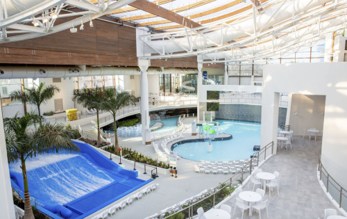 Indoor water park featuring a wave pool, water slides, and seating areas surrounded by palm trees and natural light.