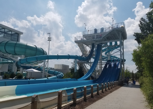 A water park featuring tall blue water slides and a sunny sky with fluffy clouds. People are enjoying the area.