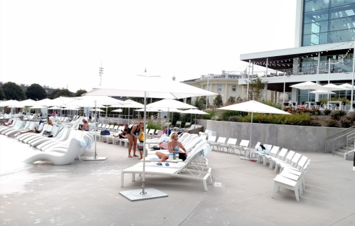 A poolside area with white lounge chairs and umbrellas, people relaxing, and a modern building in the background.