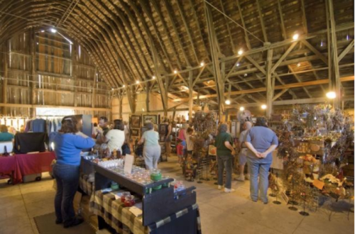 A spacious barn filled with people browsing art and crafts displayed on tables and stands. Warm, rustic atmosphere.