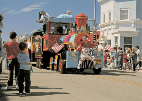A colorful train parade float with people waving, surrounded by onlookers on a sunny day.