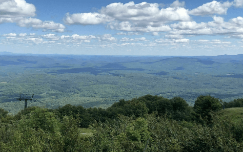 A panoramic view of lush green mountains under a blue sky with fluffy clouds, featuring a ski lift in the foreground.