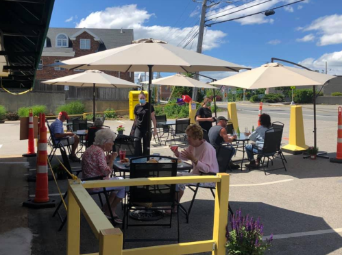 Outdoor dining scene with patrons at tables under umbrellas, a server wearing a mask, and traffic cones in the background.