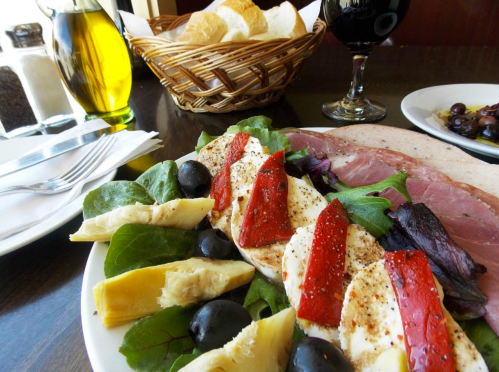 A plate of salad with mozzarella, olives, artichokes, roasted peppers, and cured meats, with bread and a drink in the background.
