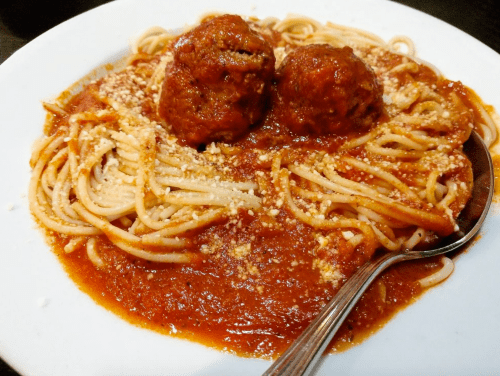 A plate of spaghetti topped with two meatballs and marinara sauce, sprinkled with grated cheese. A spoon rests beside it.