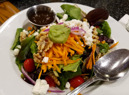 A fresh salad with mixed greens, cherry tomatoes, walnuts, shredded carrots, goat cheese, and a side of dressing.