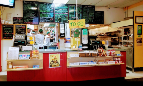 A busy restaurant counter with a menu board, staff preparing food, and a sign indicating where to order takeout.
