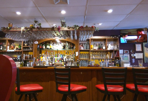 A cozy bar with a wooden counter, shelves of liquor, and red stools, featuring a TV in the background.
