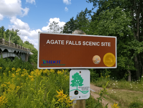 Sign for Agate Falls Scenic Site, featuring logos from MDOT and Michigan Natural Trust Fund, surrounded by greenery.