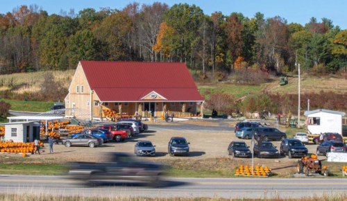 A rustic farm building with a red roof, surrounded by colorful autumn trees and parked cars, near a pumpkin patch.