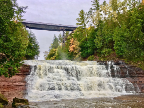 A cascading waterfall surrounded by lush greenery, with a bridge overhead and autumn foliage in the background.