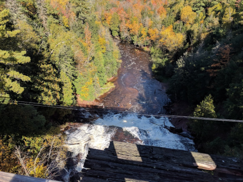 Aerial view of a river with cascading water, surrounded by vibrant autumn foliage and dense trees.