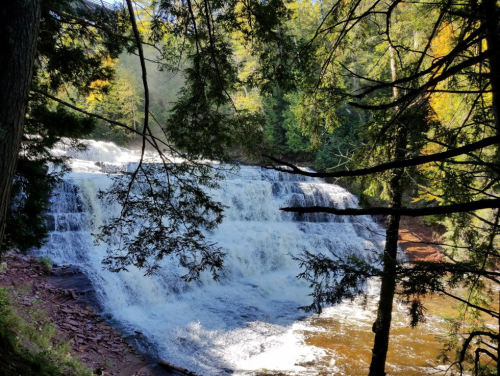 A serene waterfall cascades over rocks, surrounded by lush green trees and autumn foliage.