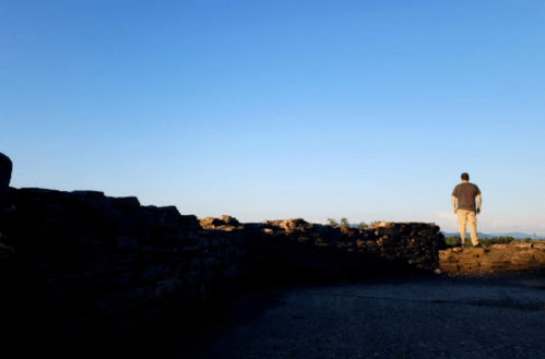 A person stands on a stone wall, gazing at a clear blue sky and distant mountains.